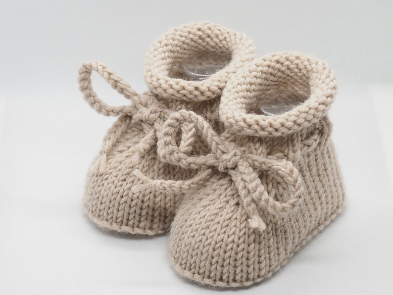 Pu&ograve; includere: Un paio di scarpine per neonati in maglia beige con un fiocco legato su ciascuna. Le scarpine sono in maglia morbida e testurizzata e sono perfette per mantenere i piedini caldi.