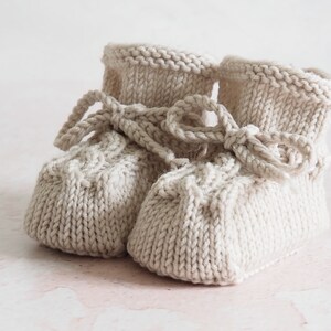 K&ouml;nnte beinhalten: Ein Paar gestrickte Babyschuhe in Cremefarbe mit einem Zopfmuster und einer Schleife.