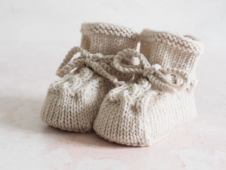 K&ouml;nnte beinhalten: Ein Paar gestrickte Babyschuhe in Cremefarbe mit einem Zopfmuster und einer Schleife.