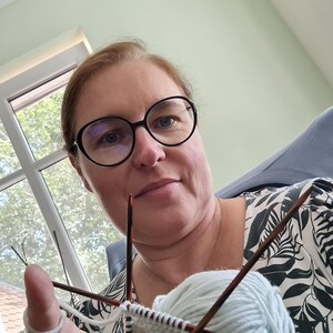 May include: A woman wearing glasses is knitting with two needles and a ball of light blue yarn. She is wearing a white and black floral shirt.