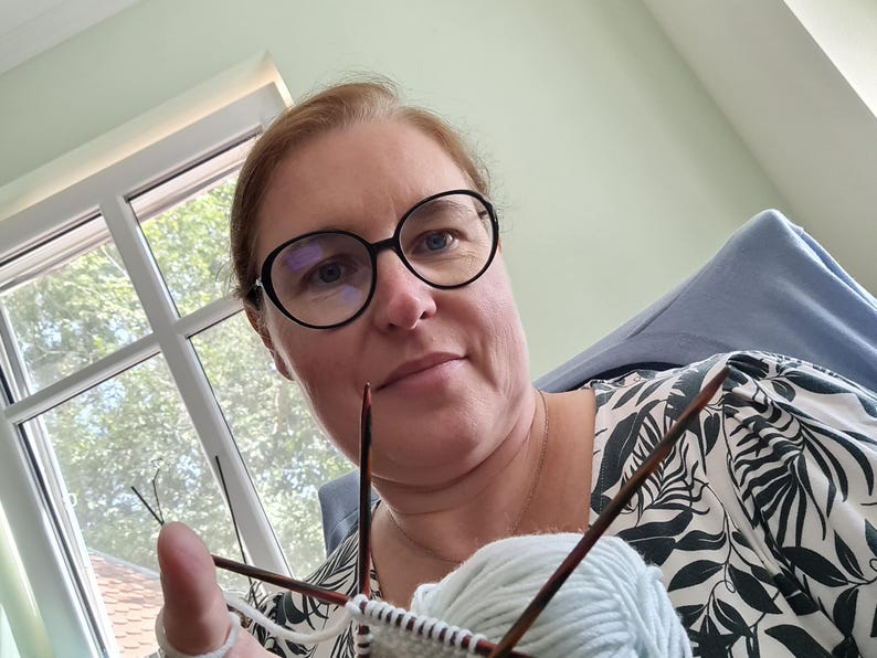 May include: A woman wearing glasses is knitting with two needles and a ball of light blue yarn. She is wearing a white and black floral shirt.
