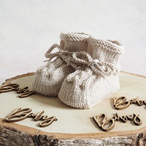 K&ouml;nnte beinhalten: Ein Paar beige gestrickte Babyschuhe mit einer Schleife auf einer Holzscheibe mit dem Wort "Baby" viermal darauf.