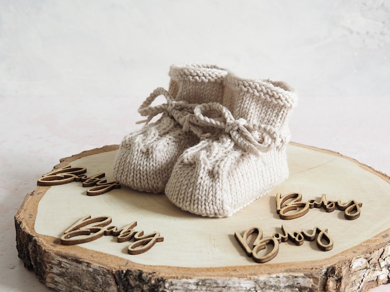 K&ouml;nnte beinhalten: Ein Paar beige gestrickte Babyschuhe mit einer Schleife auf einer Holzscheibe mit dem Wort "Baby" viermal darauf.