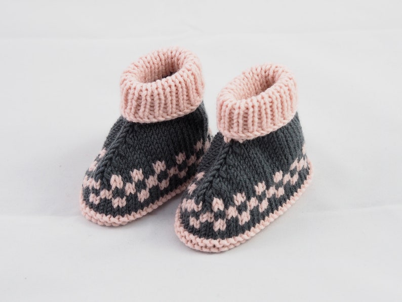 May include: A pair of knitted baby booties with a gray and pink color scheme. The booties have a pink cuff and a gray body with a pink geometric pattern.