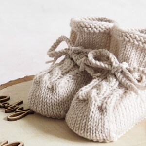 K&ouml;nnte beinhalten: Ein Paar beige gestrickte Babyschuhe mit einer Schleife. Die Schuhe liegen auf einer Holzoberfl&auml;che mit dem Wort "Baby" darauf.