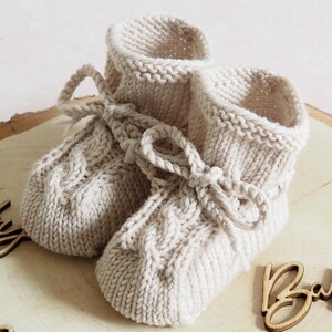 K&ouml;nnte beinhalten: Ein Paar gestrickte Babyschuhe in Cremefarbe mit einem Zopfmuster und einer Schleife. Die Schuhe liegen auf einer Holzoberfl&auml;che mit dem Wort "Baby" darauf.