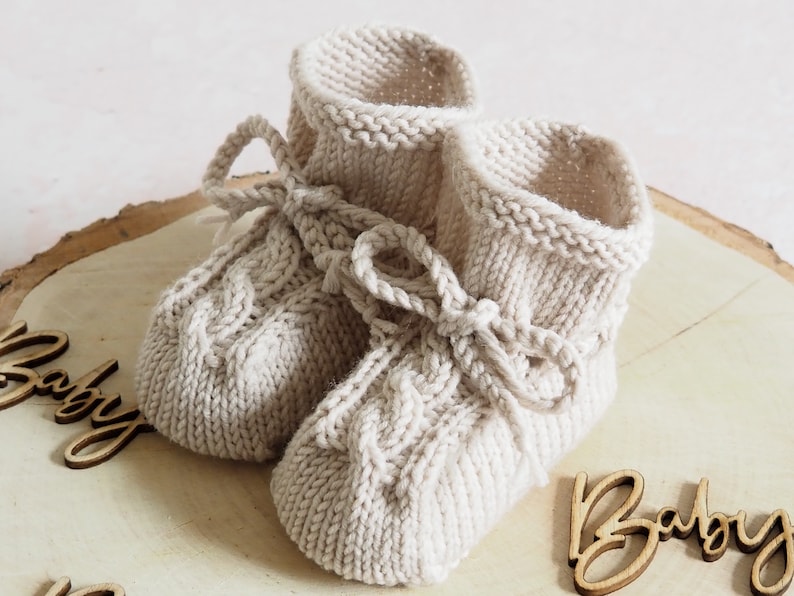 K&ouml;nnte beinhalten: Ein Paar gestrickte Babyschuhe in Cremefarbe mit einem Zopfmuster und einer Schleife. Die Schuhe liegen auf einer Holzoberfl&auml;che mit dem Wort "Baby" darauf.