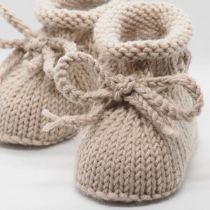 Pu&ograve; includere: Un paio di scarpine per neonati in maglia beige con un fiocco su ciascuna scarpa. Le scarpine sono realizzate in un filato morbido e testurizzato.