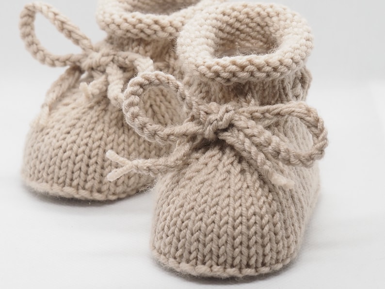 Pu&ograve; includere: Un paio di scarpine per neonati in maglia beige con un fiocco su ciascuna scarpa. Le scarpine sono realizzate in un filato morbido e testurizzato.