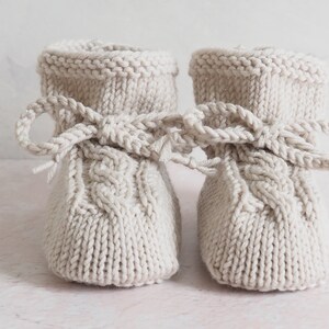 K&ouml;nnte beinhalten: Ein Paar gestrickte Babyschuhe in hellbeige mit einem Zopfmuster und einer Schleife oben.