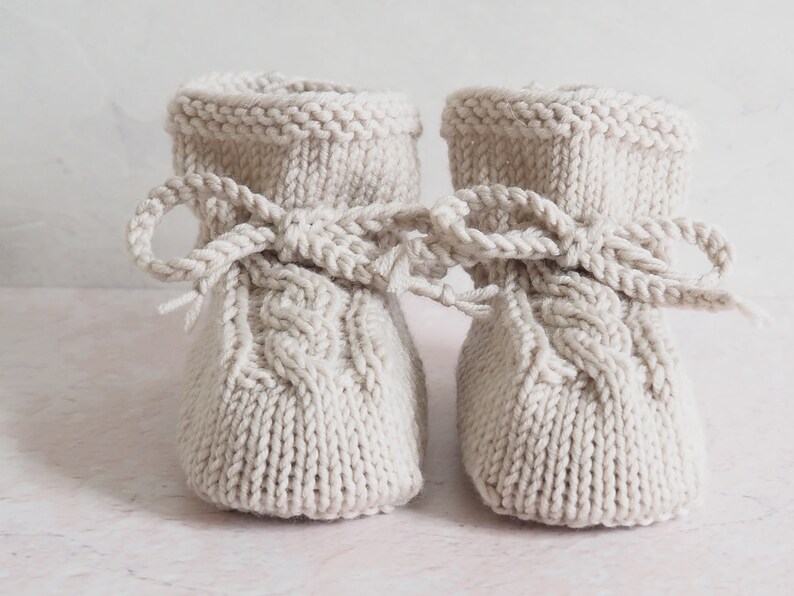 K&ouml;nnte beinhalten: Ein Paar gestrickte Babyschuhe in hellbeige mit einem Zopfmuster und einer Schleife oben.