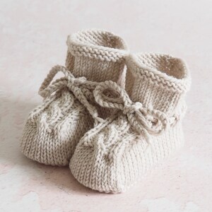K&ouml;nnte beinhalten: Ein Paar gestrickte Babyschuhe in Cremefarbe mit Schleifen-Detail.