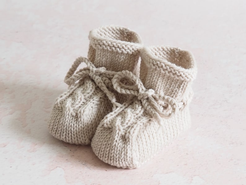 K&ouml;nnte beinhalten: Ein Paar gestrickte Babyschuhe in Cremefarbe mit Schleifen-Detail.