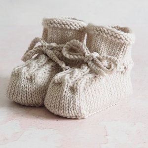K&ouml;nnte beinhalten: Ein Paar gestrickte Babyschuhe in Cremefarbe mit einer Schleife oben. Die Schuhe haben ein strukturiertes Strickmuster.