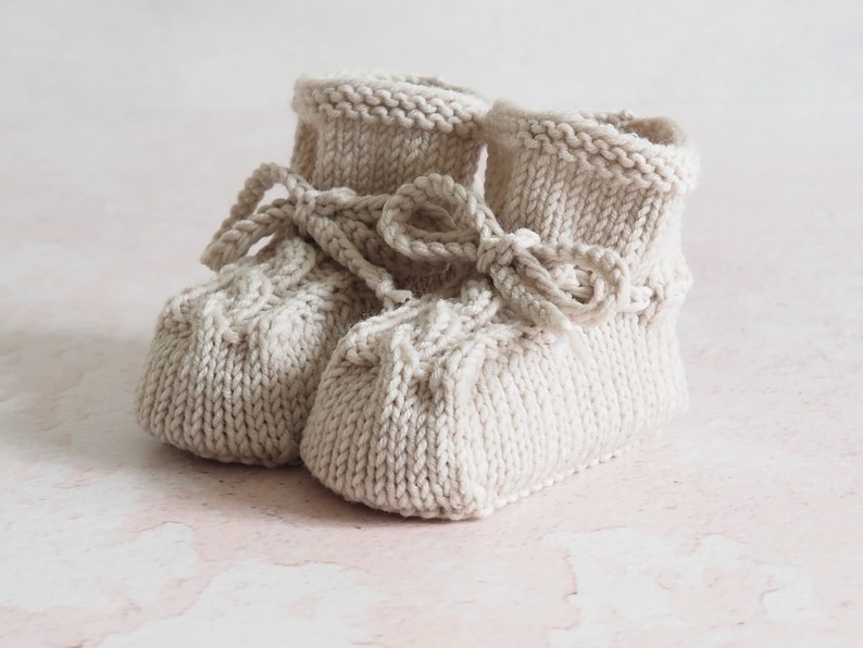 K&ouml;nnte beinhalten: Ein Paar gestrickte Babyschuhe in Cremefarbe mit einer Schleife oben. Die Schuhe haben ein strukturiertes Strickmuster.