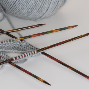 May include: A close-up of a gray knitted fabric on four colorful wooden knitting needles. The needles are crossed over each other and the yarn is wrapped around them.