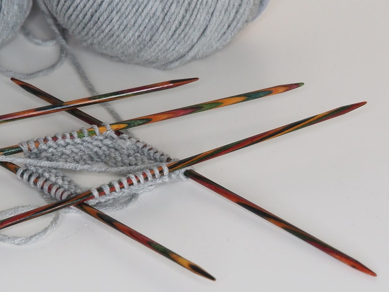 May include: A close-up of a gray knitted fabric on four colorful wooden knitting needles. The needles are crossed over each other and the yarn is wrapped around them.