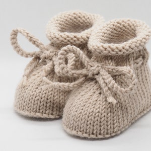 Pu&ograve; includere: Un paio di scarpine per neonati in maglia beige con un fiocco legato su ogni scarpa.