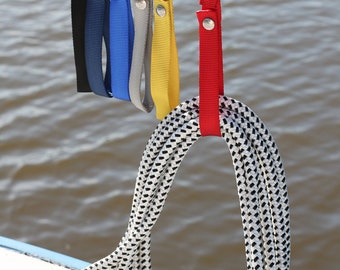 May include: A close-up of a coiled white and black rope, held by a red strap. Several colored straps in black, navy, blue, gray, and yellow are visible above. The straps have metal snap closures. The background is a blurred view of water.