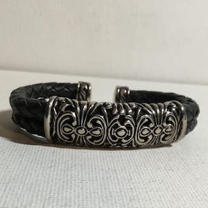 May include: A black braided leather bracelet featuring a central silver-toned metal component with intricate embossed scrollwork and floral-inspired patterns. The cuff-style bracelet has an open design with polished metal end caps.