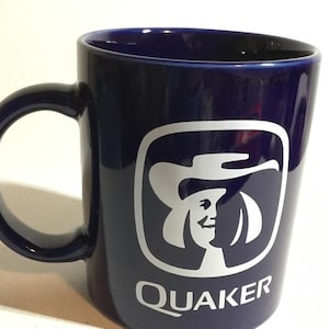 May include: A navy blue ceramic mug with a large handle. The mug features the Quaker Oats logo in white, including the image of a woman in a hat, with the word "Quaker" below.
