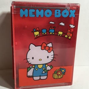 Puede incluir: Una caja de notas de plástico rojo con un diseño de Hello Kitty. La caja tiene las palabras "MEMO BOX" en letras azules. Debajo, Hello Kitty está representada con un mono azul, sosteniendo un objeto verde y una cesta de flores.