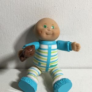 May include: A vintage Cabbage Patch Kids doll in a seated position. The doll has a bald head, green eyes, and rosy cheeks. It wears a blue, white, and green striped onesie with matching blue booties. The doll is holding a brown teddy bear.