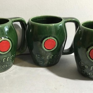 Könnte beinhalten: Drei grüne Keramikbecher mit roten kreisförmigen Akzenten und den Worten "Coffee D.O.I." auf der Vorderseite.