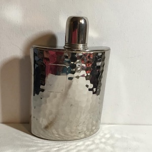 May include: A silver flask with a hammered finish and a rounded rectangular form. It features a small, cylindrical cap. The surface reflects light, giving a textured appearance. The flask is likely made of metal.
