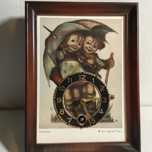 May include: A framed clock featuring a vintage illustration of two children under an umbrella. The clock face is black with gold hands and numerals. The frame is dark brown, and the image has the text "Hummel" and "Ars sacra 9743".