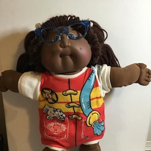 以下が含まれることがあります： 茶色の髪と肌を持つキャベツパッチキッズのドール。青い眼鏡をかけ、赤、黄、青のライフガードの服を着ています。犬のグラフィックと「キャベツパッチキッズ」の文字が書かれています。