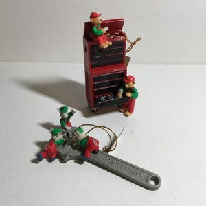 Peut inclure: Une décoration de boîte à outils Craftsman rouge avec quatre petites figurines de mécaniciens. La boîte à outils est munie d'une clé à molette argentée fixée au fond. Les figurines portent des tenues rouges et vertes.