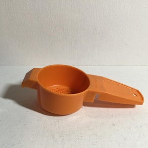 May include: Orange plastic kitchen utensil with a round, perforated bowl and a long handle. The handle has a small hole at the end. The tool is for straining or sifting ingredients.
