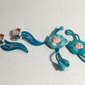 May include: Two blue plastic hair clips with a flower design and a cartoon face. Two blue plastic hair clips with a cartoon Hello Kitty design.