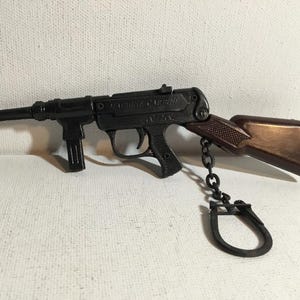 Op de afbeelding: Zwart speelgoed machinegeweer met een bruine kolf en een metalen ketting bevestigd aan een sleutelhanger. De woorden "MACHINE CARBINE" zijn zichtbaar op de zijkant van het geweer.