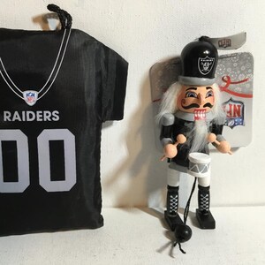 May include: Black Raiders jersey-shaped ornament with the number 00 and the word RAIDERS in white. A wooden nutcracker in a black and silver uniform with a drum and drumsticks is also present. The nutcracker has a white beard and mustache.