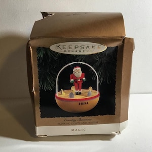 Könnte beinhalten: Eine braune und goldene Schachtel mit einem Weihnachtsbaumschmuck. Der Schmuck ist eine Holzfigur des Weihnachtsmanns in einem roten Anzug, die auf einer Plattform mit blinkenden Lichtern steht. Die Schachtel trägt die Aufschrift "Keepsake Ornament" und "Country Showtime Blinking Lights and Motion Magic".