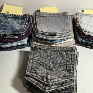 Puede incluir: Montones de bolsillos de mezclilla en varios tonos de azul, gris y morado. Los bolsillos están cuidadosamente doblados y dispuestos en pilas separadas. Algunos bolsillos tienen costuras visibles y el diseño clásico de Levi's. Cada pila tiene una nota adhesiva amarilla encima.