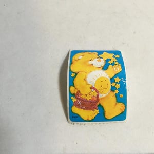 May include: A yellow Care Bear with a blue star on its chest and a basket of flowers in its hands. The bear is standing on a blue background with white stars.