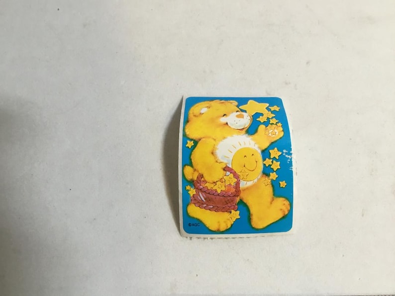May include: A yellow Care Bear with a blue star on its chest and a basket of flowers in its hands. The bear is standing on a blue background with white stars.