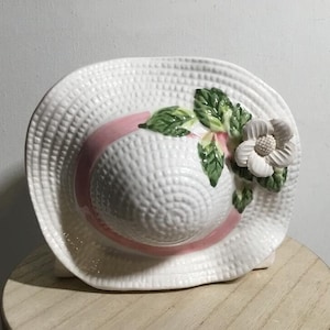 May include: White ceramic planter shaped like a straw hat with a pink ribbon and a white flower with green leaves.