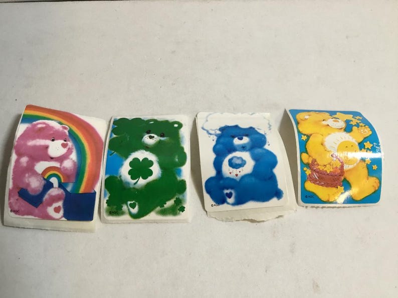 May include: Four Care Bear stickers featuring different Care Bears. The stickers are on a white background. The Care Bears are all smiling and have different colored fur. The Care Bears are all wearing different outfits. The stickers are all rectangular in shape.