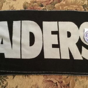 Può includere: Sciarpa nera e bianca dei Raiders con un'etichetta bianca di prodotto con licenza NFL. La sciarpa ha un bordo con frange.