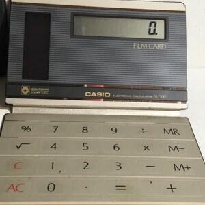 May include: A vintage Casio SL-300 electronic calculator with a gray and black design. The calculator displays "0." on its screen and features a solar cell. The keyboard has white keys with black numbers and symbols.