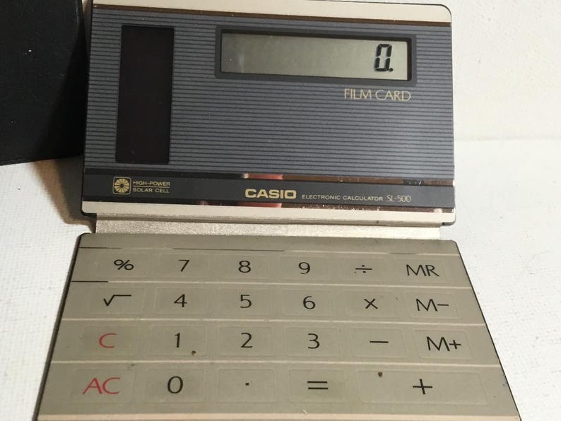 May include: A vintage Casio SL-300 electronic calculator with a gray and black design. The calculator displays "0." on its screen and features a solar cell. The keyboard has white keys with black numbers and symbols.