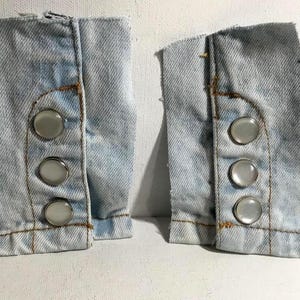 May include: Two light blue denim cuffs with three white plastic buttons each. The buttons are sewn onto the cuffs.