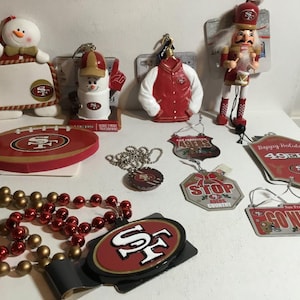 Könnte beinhalten: Eine Sammlung von Ornamenten und Dekorationen zum Thema San Francisco 49ers. Enthält einen fußballförmigen Schmuck, einen Schneemann, einen Nussknacker, einen Jackenanhänger und Perlenketten. Das Farbschema ist rot, gold und weiß.