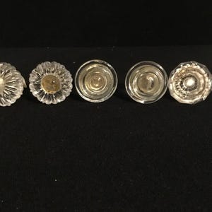 May include: Five clear glass cabinet knobs arranged in a horizontal row. The knobs feature varied designs, including fluted, petal-like, and smooth circular patterns with reflective surfaces, showcasing different textures and shapes in clear glass.