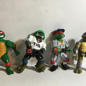 Puede incluir: Cuatro figuras de acción de las Tortugas Ninja. Cada figura es verde con máscaras de diferentes colores, incluyendo rojo, verde y morado. Una figura lleva un uniforme de béisbol, y otra una camiseta con "THL".