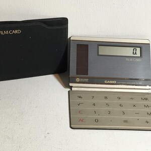 May include: A vintage Casio Film Card calculator with a gray and silver design, featuring a numeric keypad and a digital display showing "0". The calculator is open, next to a black case with "CASIO FILM CARD" printed in gold.